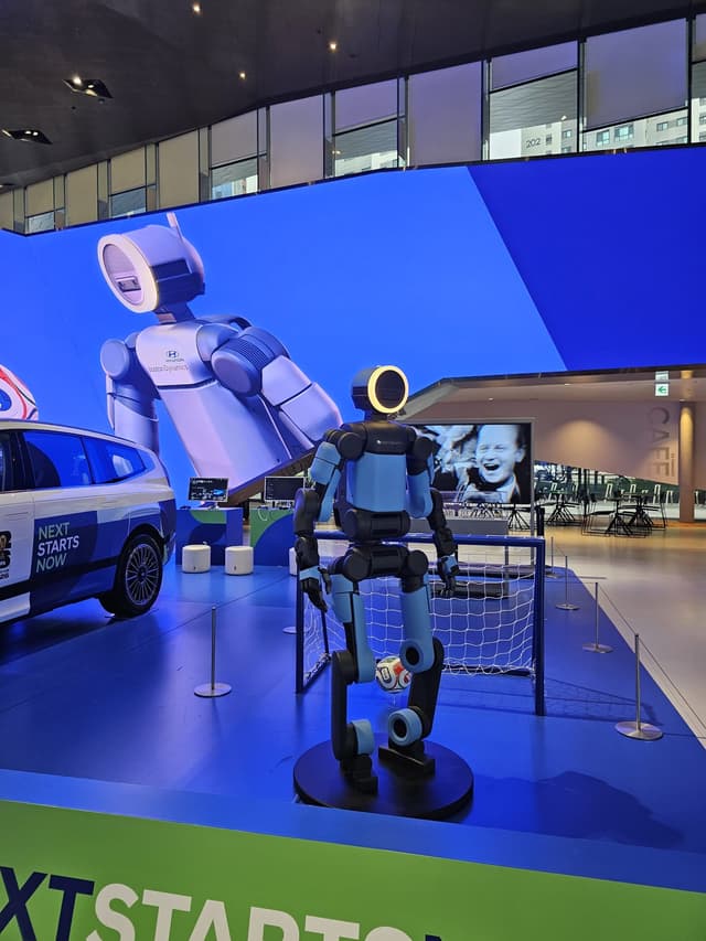 Boston Dynamics Atlas humanoid robot standing on a display pedestal in front of a small football goal, with a large image of Atlas on the screen behind it.