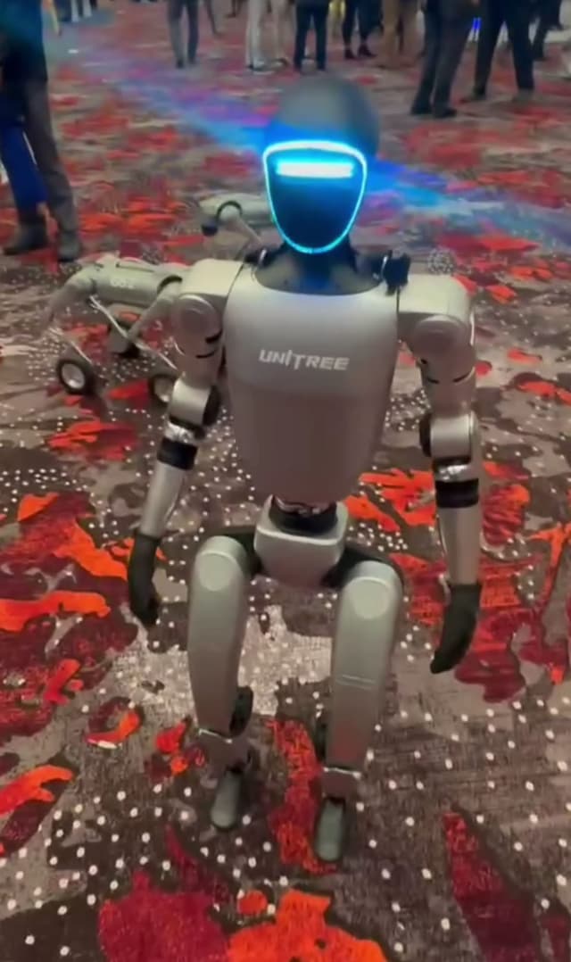 Unitree G1 humanoid robot standing on a patterned exhibition carpet, facing the camera, its face panel lit blue and the Unitree logo on its chest.