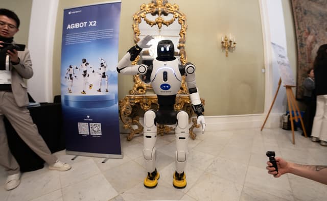 AGIBOT X2 humanoid robot standing and waving beside an AGIBOT X2 banner in an ornate room, with visitors filming it.